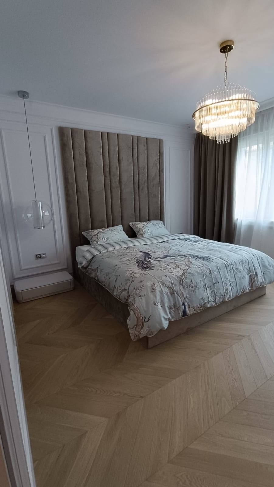 Chambre avec haute tête de lit capitonnée en velours, lustre en cristal, boiseries murales et parquet à chevrons